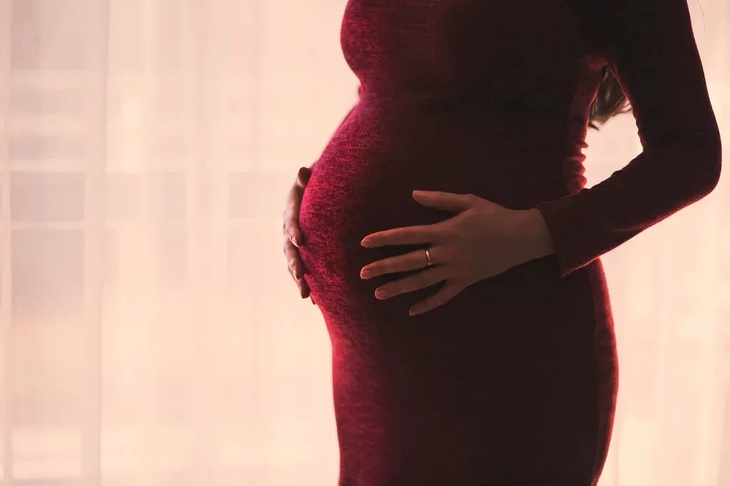 Future maman en robe, la main posée sur son ventre, devant une fenêtre lumineuse